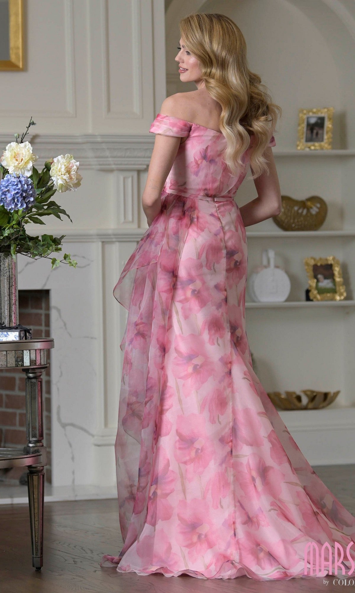A woman with long wavy hair wears the Marsoni MV1394 long formal dress, standing indoors near a fireplace and decorative shelves.